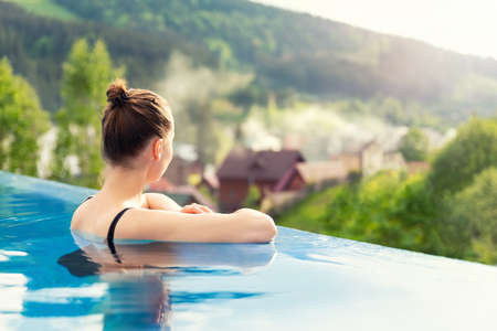 back view young adult female person enjoy relax in infinity edge luxury outdoor swimming pool looking on fog hill green mountains in warm sunset light. Alpine welness travel vacation tourism conceptの写真素材