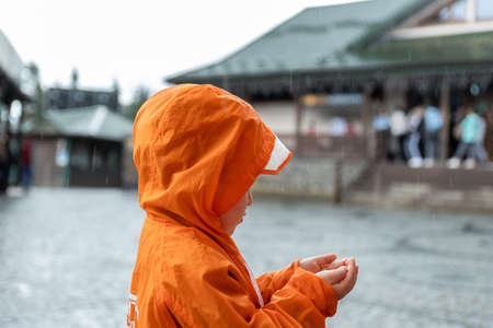Portrait cute little lonely caucasian kid boy in bright orange waterproof raincoat stand under rain and enjoy catching drop in hands on european city street outside. Autumn weather outdoor walkingの写真素材