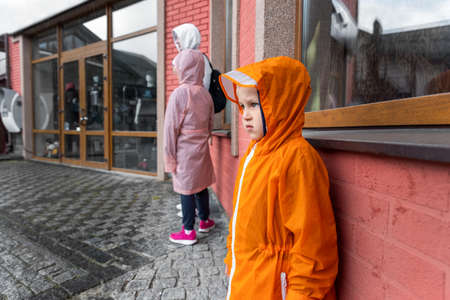 Portrait cute little lonely caucasian kid boy in bright orange waterproof raincoat stand under rain against brick wall building european city street with mother and sister outside. Autumn fall moodの写真素材