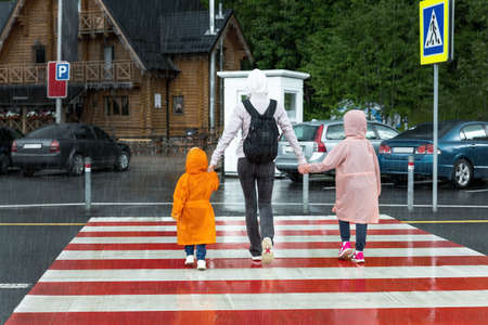 Back view of young adult mother hold daughter and son hand crossing zebra crosswalk road hiking at mountain village during shower rain at atumn day. People walking crossroad. Children traffic safetyの写真素材