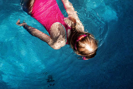 Above top view of little cute kid girl in sport pink swimsuit and goggles diving underwater in clear blue water of indoor swimming pool at dark evening time. Children watersport healthy activitiesの写真素材