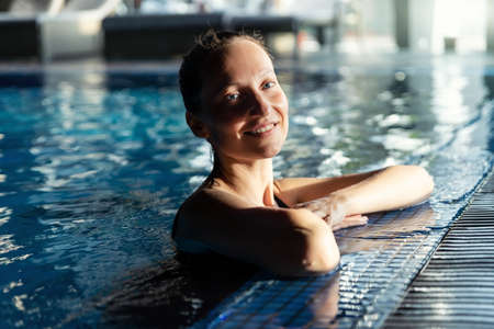 Portrait of young adult caucasian beautiful female model in sport black swimsuit in clear blue water edge indoor swimming pool at dark evening time. Relax harmony and wellness spa therapyの写真素材