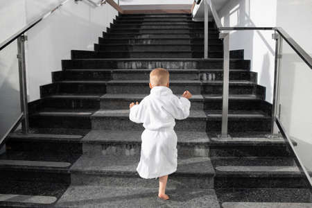 Back view of cute funny caucasian blond kid boy wearing white pool spa bathrobe walking upstairs on big marble staircase. Alone small child meet challenge and purpose. Rich luxury lifestyle conceptの写真素材
