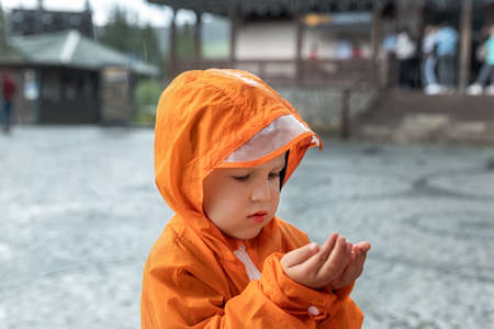 Portrait cute little lonely caucasian kid boy in bright orange waterproof raincoat stand under rain and enjoy catching drop in hands on european city street outside. Autumn weather outdoor walkingの写真素材