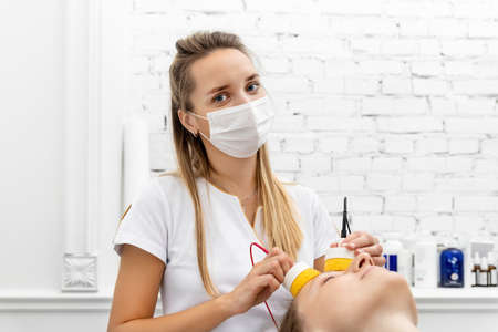 Young adult attractive professional beautician doctor portrait in protective face mask do facial care and treatment procedure to female client. Health safety therapy.の写真素材