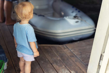 Back view of cute little caucasian toddler boy stand alone in ship garage wooden floor dock and looking on inflatable boat dreaming of sailing by lake or pond river. Summer outdoor activityの写真素材