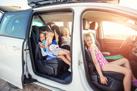 Group of four cute adorable little happy caucasian children enjoy having fun sit in minivan going to sea beach road trip on hot summer day. Kids in swimsuit in car with side open door against sunの写真素材
