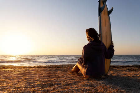 Back view young adult slim sporty female surfer girl with surfboard sitting on sand at ocean coast wave against warm sunrise or sunset sun. Sport healthy carefree freedom lifestyle vacation conceptの写真素材