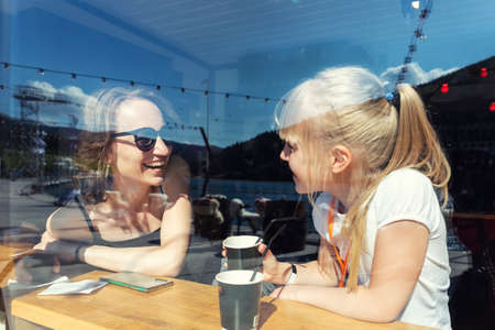 Through window view of young adult happy mom enjoy having fun talking, drink hot chocolate laugh with cute little daughter sitting at cafe restaurant on sunny day. Mother and child friendship conceptの写真素材