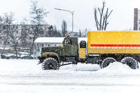 Scenic view snow covered city street with big old powerful assistance tow service truck on road snow drift. Snowfall danger blizzard bad winter weather. Urban cold foggy day snowstrom town backgroundの写真素材