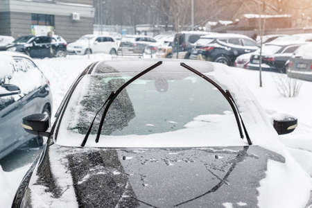 Close-up detail view of black modern car with raised up windshield wiper blades to prevent icing in cold frost winter day. Vehicle windscreen covered by whote snow parked on city street outdoorsの写真素材
