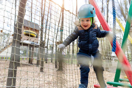 Portrait happy brave courage little toddler child boy wear safety equipment helmet enjoy passing obstacle course forest rope adventure park on cold winter day. Active outside leisure amusement campの写真素材
