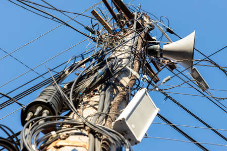 Chaos of many different data cables and network wires messy tangled hanged on concrete pillar pole on urban city street against clear blue sky on day time. Announcement speaker mounted on columnの写真素材