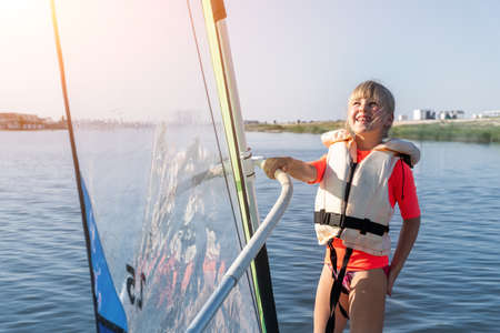 Cute little caucasian blond child girl enjoy having fun learning to surf on winsurfing board at freshwater pond lake or river hot sunny day. Summer outdoors child healthy sport recreation activitiesの写真素材