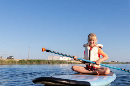 Cute little caucasian blond child girl enjoy having fun sitting on sup board surfing at freshwater...