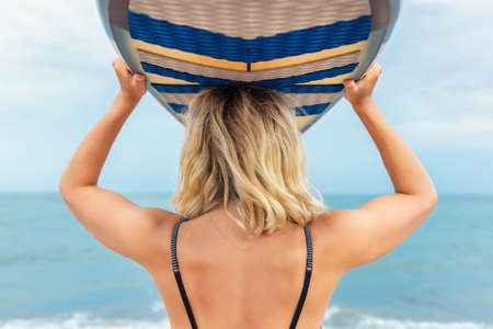 Back view young adult slim sporty female surfer girl enjoy having fun carrying surfboard head against ocean coast wave clear water sunny day. Sport healthy carefree freedom lifestyle vacation conceptの写真素材