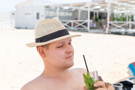Portrait of young adult caucasian man wearing hat enjoy having fun drinking fresh mojito cocktail and sunbathing on sand beach on hot summer day. Seaside or ocean leisure vacation conceptの写真素材