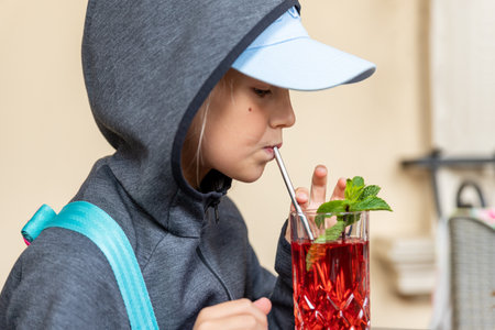 Profile portrait view of cute preteen little kid girl in grey hoodie enjoy drinkong healthy berry lemonade juice compote cocktail walking outdoors. Thirsty child drink beverage outsideの写真素材