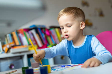 Portrait of cute little blond kid boy enjoy have fun painting with watercolor and brush bright abstract image on paper at home or kindergarten class. Imagination development children play activityの写真素材
