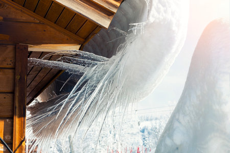 Close-up detail view of melting snow icicles on wooden alpine hut roof at windy mountain. Beatiful croocked big icicles on cold frosty sunny winter dayの写真素材