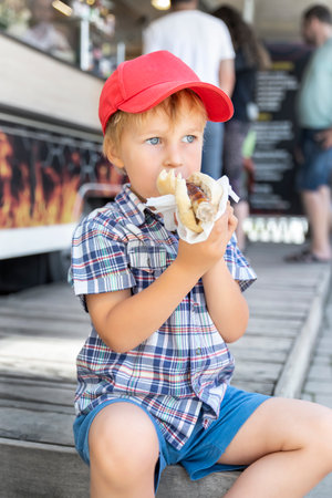 Cute adorable pensive little boy kid enjoy eating hot dog sausage in bread near street cafe stall outdoors. Child healthy eating lunch hotdog. Junk food and fast food unhealthy snack conceptの写真素材