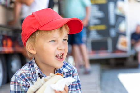 Cute adorable little happy smiling boy kid enjoy eating hot dog sausage in bread near street cafe stall outdoors. Child healthy eating lunch hotdog. Junk food and fast food unhealthy snack conceptの写真素材
