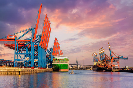 Scenic front giant cargo container ship loading Hamburg city port harbour seaport cranes warm dramatic sky evening sunset light. Global commercial trade freight charter shipping logistics backgroundの写真素材