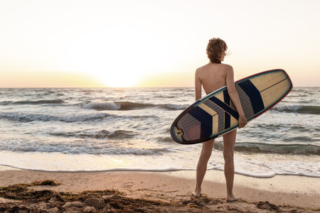 Back view young adult slim sporty female surfer girl with surfboard silhouette stand at ocean coast wave against warm sunrise or sunset sun. Sport healthy carefree freedom lifestyle vacation conceptの写真素材