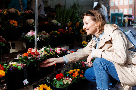 Young adult joyful woman kneels to choose buy colorful potted flowers at outdoor market stall. Sale of blooming plants in pots at botanical street German store on sunny spring dayの写真素材