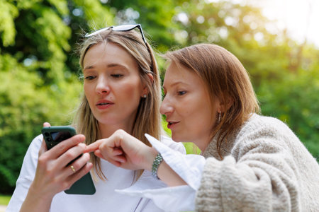 Two young adult happy smiling woman friends portrait using mobile phone smartphone outdoors. Female...