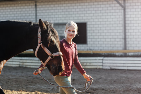 Young little teenager kid girl leading chestnut horse through stable ranch yard. Horse riding school exercise.  Cute little beginner blond girl kid near beautiful brown horse. Equine hobby or workの写真素材