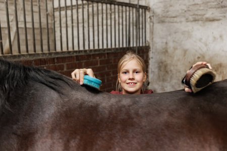 Young little teenager kid girl cleaning grooming chestnut horse back hair brush tool horse at stable ranch. Horse ride school farm life. Cute little blond girl kid care brown horse. Equine hobby workの写真素材