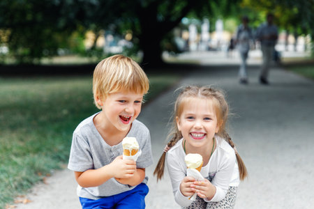 Portrait two cute cheerful children enjoy ice cream together at nature city park path on hot summer sunny day. Funny kids eat sweet vanilla icecream walking family outdoorsの写真素材