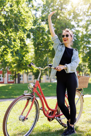 Portrait young adult pretty woman vintage retro bike and casual outfit waving hand green city park bright sunny summer day. Happy beautiful female person cycling outdoor. Healthy lifestyleの写真素材