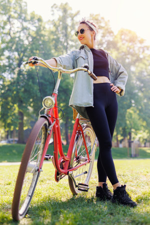 Portrait young adult pretty woman with red vintage retro bike and casual outfit green city park bright sunny summer day. Happy beautiful female person cycling outdoors. Healthy lifestyleの写真素材