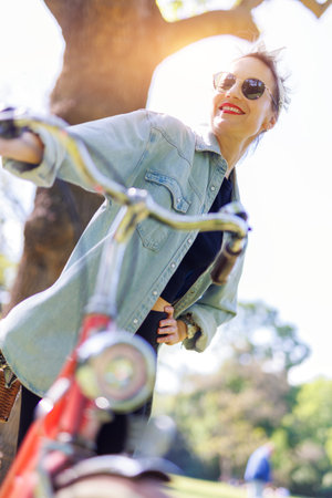 Portrait young adult pretty woman with red vintage retro bike and pin-up casual outfit green city park bright sunny summer day. Happy beautiful female person cycling outdoors. Healthy lifestyleの写真素材