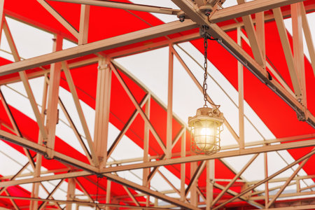 Industrial-style lantern hangs from beige steel trusses under a vibrant red and white striped canopy. Geometric structure and warm light create contrast between utility and visual boldnessの写真素材