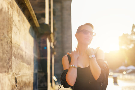 Portrait young adult elegant woman black dress and sunglasses smiles confidently in golden sunlight warm sunset backlit light against historic cathedral building. Female person walking city streetの写真素材
