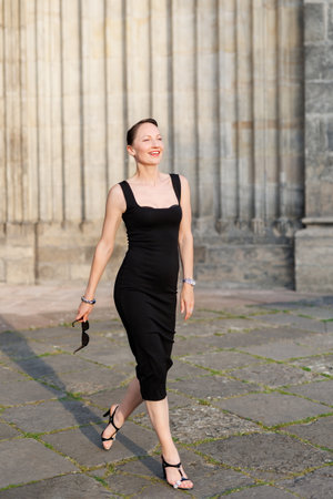 Portrait young adult elegant woman black dress and sunglasses smiles confidently in golden sunlight warm sunset backlit light against historic cathedral building. Female person walking city streetの写真素材