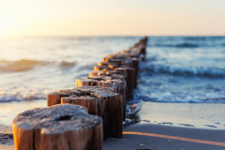 Wooden breakwater stretches into blue sea under soft evening light. Gentle waves touch sandy shore...