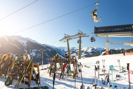 Sunny winter day on alpine slope with skis and snowboards resting near gondola station. Bright sunlight, blue sky, and mountain panorama create atmosphere of adventure, freedom, and relaxationの写真素材