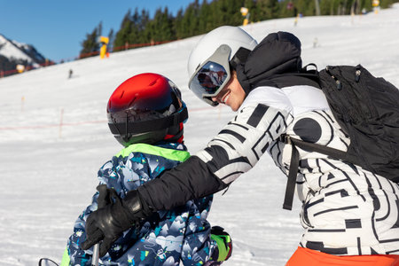 Mother teaching child to ski on sunny mountain slope, holding and guiding for balance and confidence. Warm family moment of encouragement and teamwork in bright winter landscape full of joyの写真素材