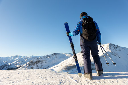 Skier stands on snowy mountain ridge facing bright horizon, ready for descent and challenge. Scene captures spirit of freedom, determination and strength in high alpine wildernessの写真素材