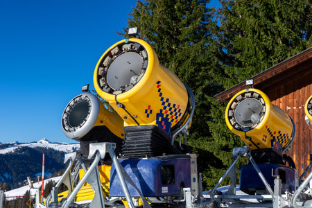 Operating artificial snow cannon near piste making snowy powder. Ski lift ropeway on highland alpine mountain winter resort on bright sunny day. Downhill slopes with people enjoying sport activitiesの写真素材
