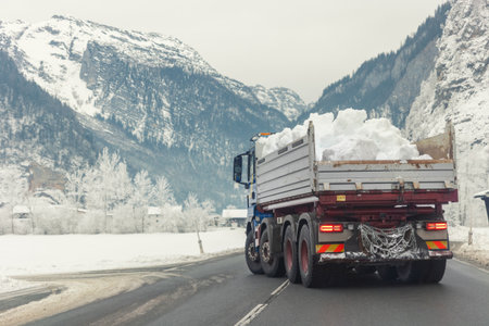 Big dump truck fully loaded with snow driving through mountain alpine road at european Alps landscape. Heavy machinery snow removal. Municipal services cleaning and maintenance town roadsの写真素材