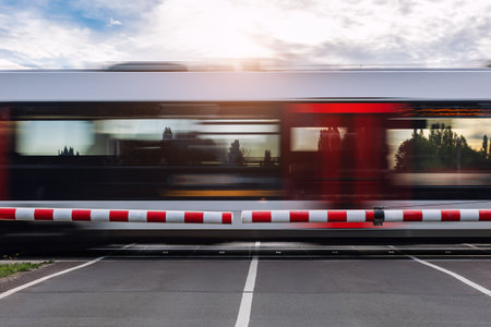 Fast modern regional commute train rushes past closed railway crossing under cloudy sky  sunlight sunset evening. Motion blur rush hour speed dynamic atmosphere  urban transportation commute sceneの写真素材