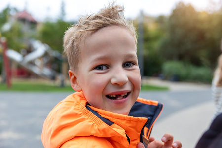 Portrait happy smiling boy with missing tooth wearing bright orange jacket at playground. Joyful expression and sunlight in background create cheerful childhood mood full of energy and spontaneityの写真素材