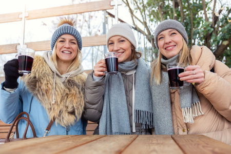 Portrait three pretty young adult woman friends enjoy laughing drinking hot mulled wine at Christmas market outdoors cold winter day. People having fun celebrate new year time street cafe outsideの写真素材