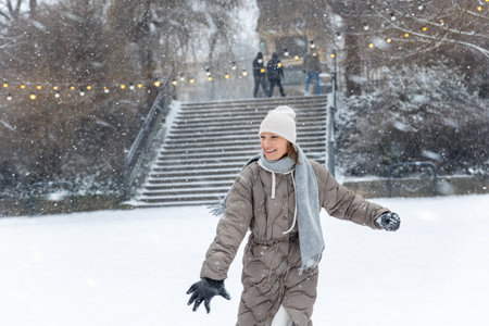 Portrait young adult smiling woman wear long quilted coat white hat enjoy play snowball snowy day outdoors. Snowflakes fall she moves playfully in front of stairs decorated with glowing winter lightsの写真素材