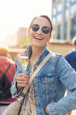 Portrait young adult pretty happy woman woman hold sparkling wine prosecco drink in lively street setting. Denim jacket floral dress sunglasses create vibrant atmosphere filled with festive energyの写真素材